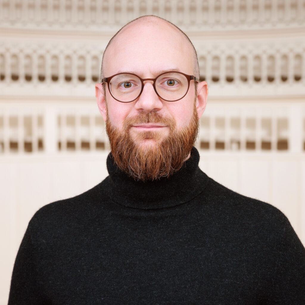 Brustporträt von Felix Sattler, Foto von Philip Plum. Ein weißer Mann mittleren Alters mit Brille, Glatze und Bart, in schwarzem Rollkragenpullover, im Auditorium des Tieranatomischen Theaters.